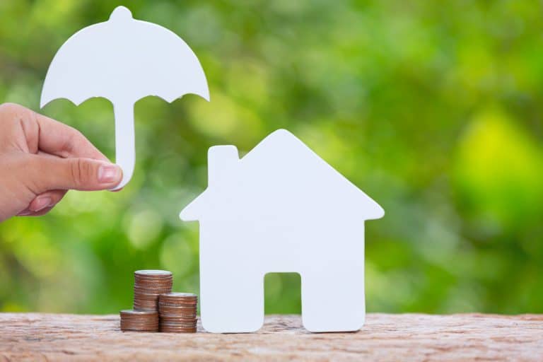 Personne tenant une petite parapluie en papier au dessus d'une maison et de pièces de monnaie, représentant une assurance habitation